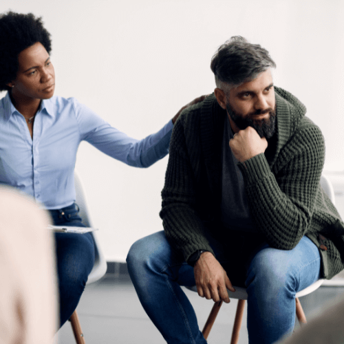 Man in rehab group therapy looking away from participants as his therapist looks at him with her hand on his shoulder